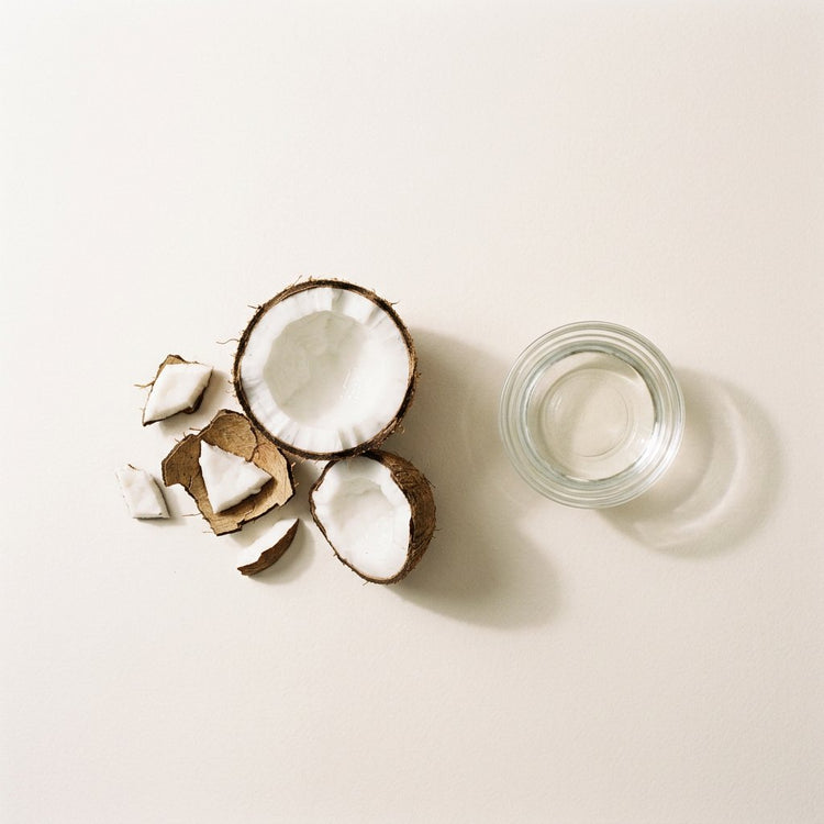 Virgin Coconut: a coconut cracked open to show its white flesh, beside a small glass dish of the oil.