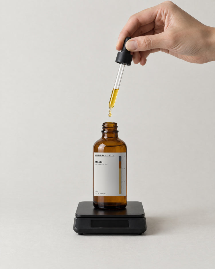 A hand releasing a drop of oil from a pipette into an amber bottle standing on a small black scale.