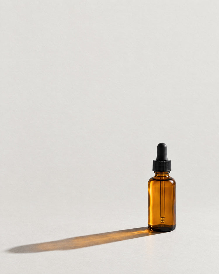 An amber glass dropper bottle on pale paper, backlit, casting one long amber shadow.